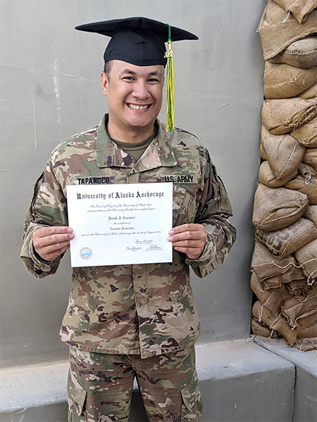 Deployed student in military uniform with grad cap and tassel on, posing with diploma.