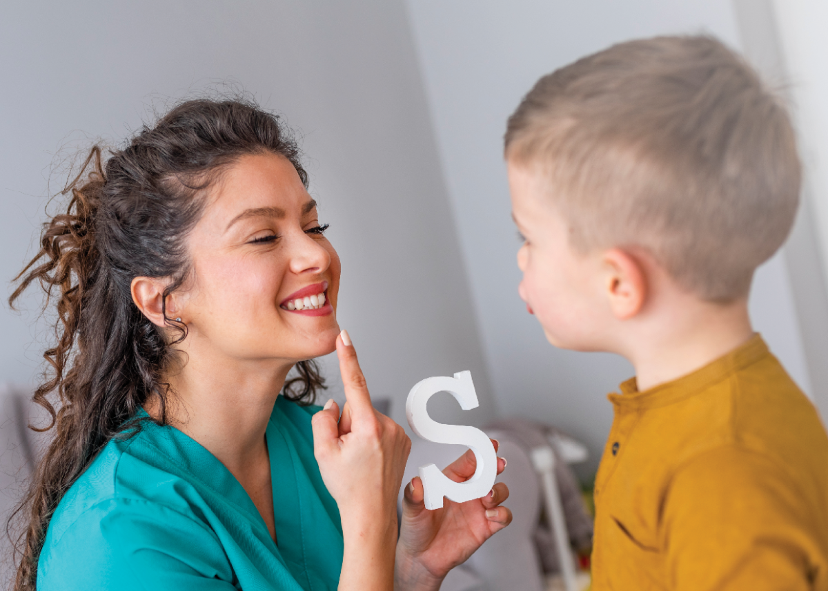 A speech language pathologist helping a child with pronunciation, pointing to their mouth, holding the letter "S."