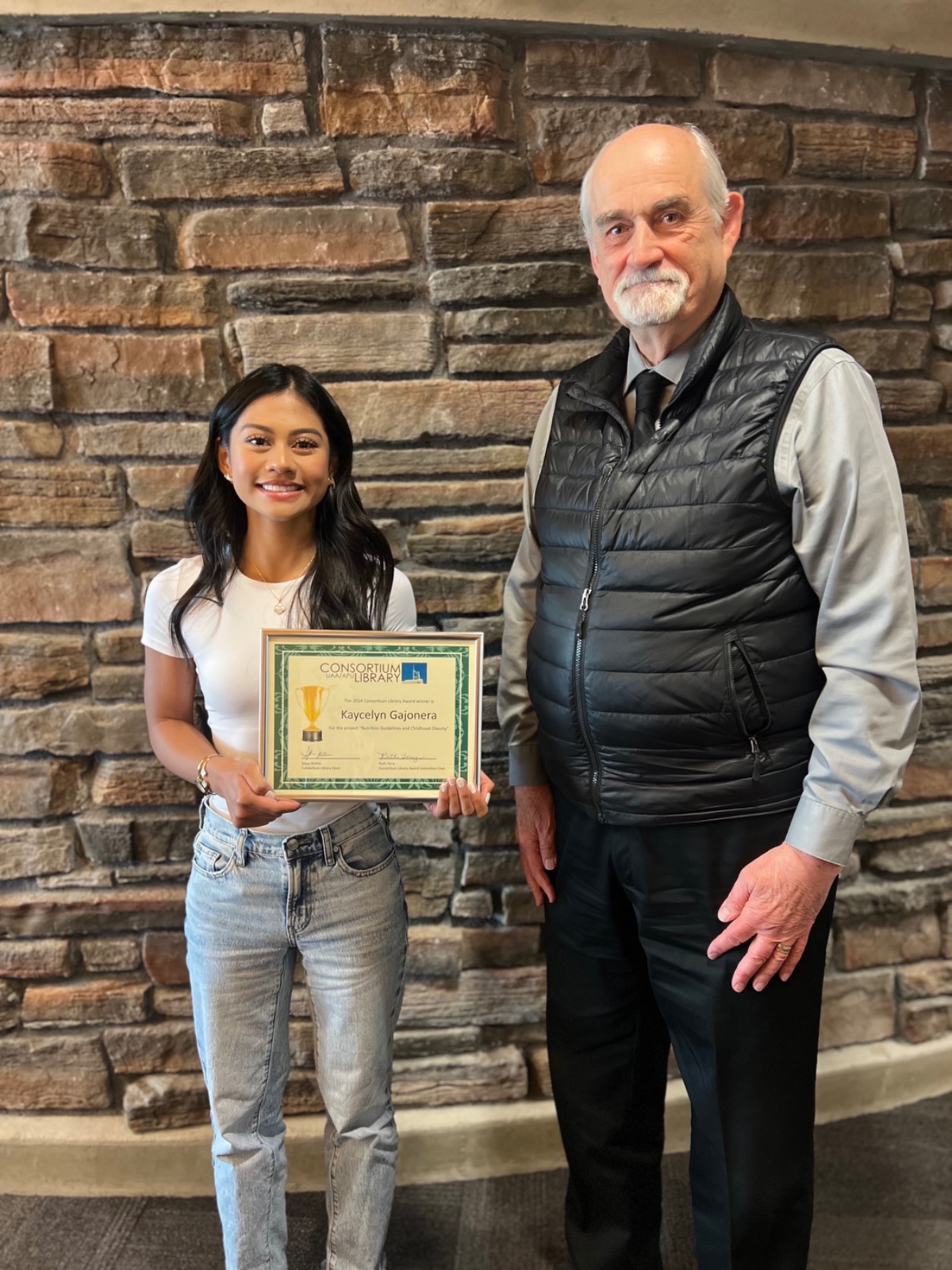 Kaycelyn Gajonera stands with Library Dean Steve Rollins as she accepts the 2024 UAA Consortium Library Award.