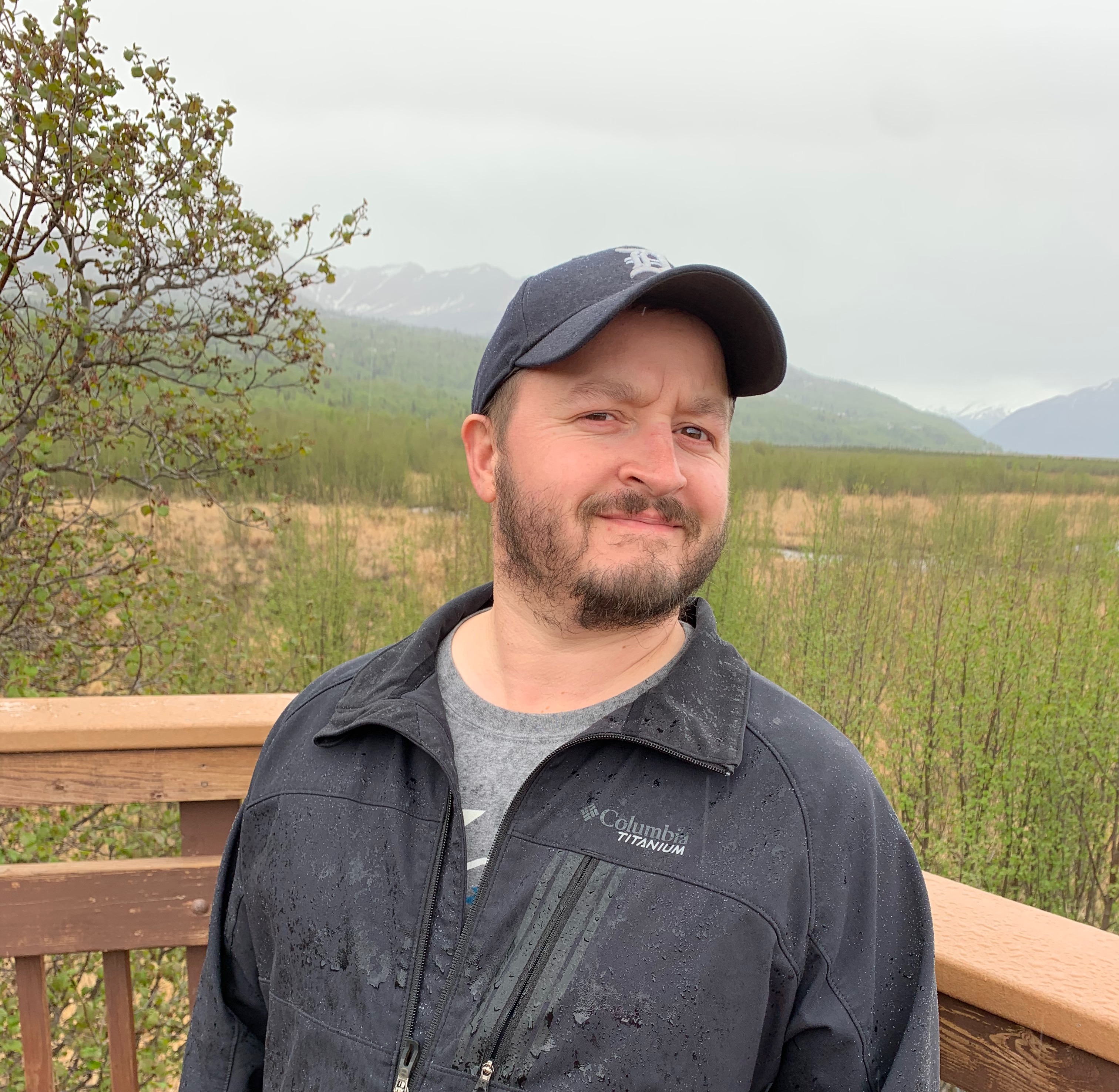 A headshot of Zebadiah wearing a baseball cap and standing on a deck with mountains in the background